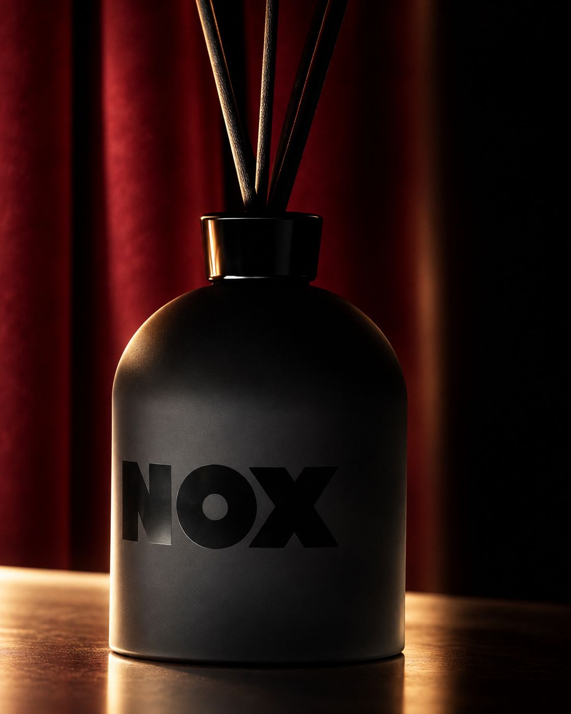 Το NOX Reed Diffuser, ματ μαύρο μπουκάλι με σκούρα στικ μπροστά σε κόκκινη βελούδινη αυλαία